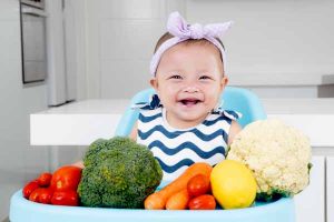 Adorable baby girl smiling and sitting on baby chair with vegetables in the kitchen